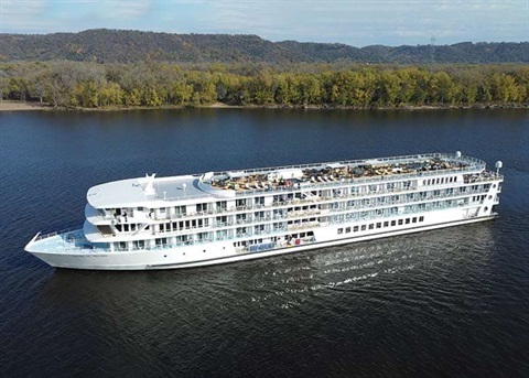 A large white cruise ship travels down a wide, dark river with tree-covered hills in the background under a partly cloudy sky.