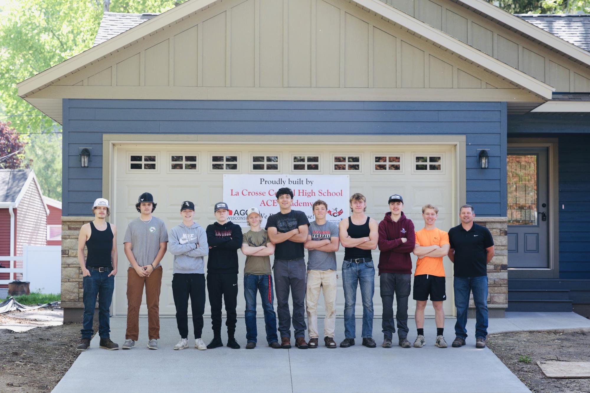 A group of students and an instructor stand in front of a newly built house with a banner that reads "Proudly built by La Crosse County High School ACE Academy"