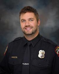 Adult male wearing navy-colored police uniform posed for a professional portrait against a dark background.
