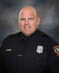 Adult male wearing navy-colored police uniform posed for a professional portrait against a dark background.