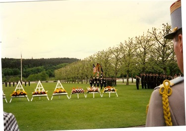 Man in military dress viewing rows of memorial flowers on white easel stands in a cemetery