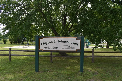 Sign for Clayton E. Johnson Park, established in 1995, with playground equipment visible behind a wooden fence.