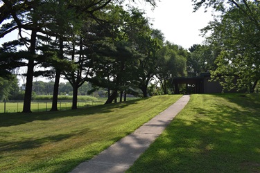 A paved walking path curves uphill towards a modern-style building with a dark roof, surrounded by lush green grass and mature trees on a sunny day.