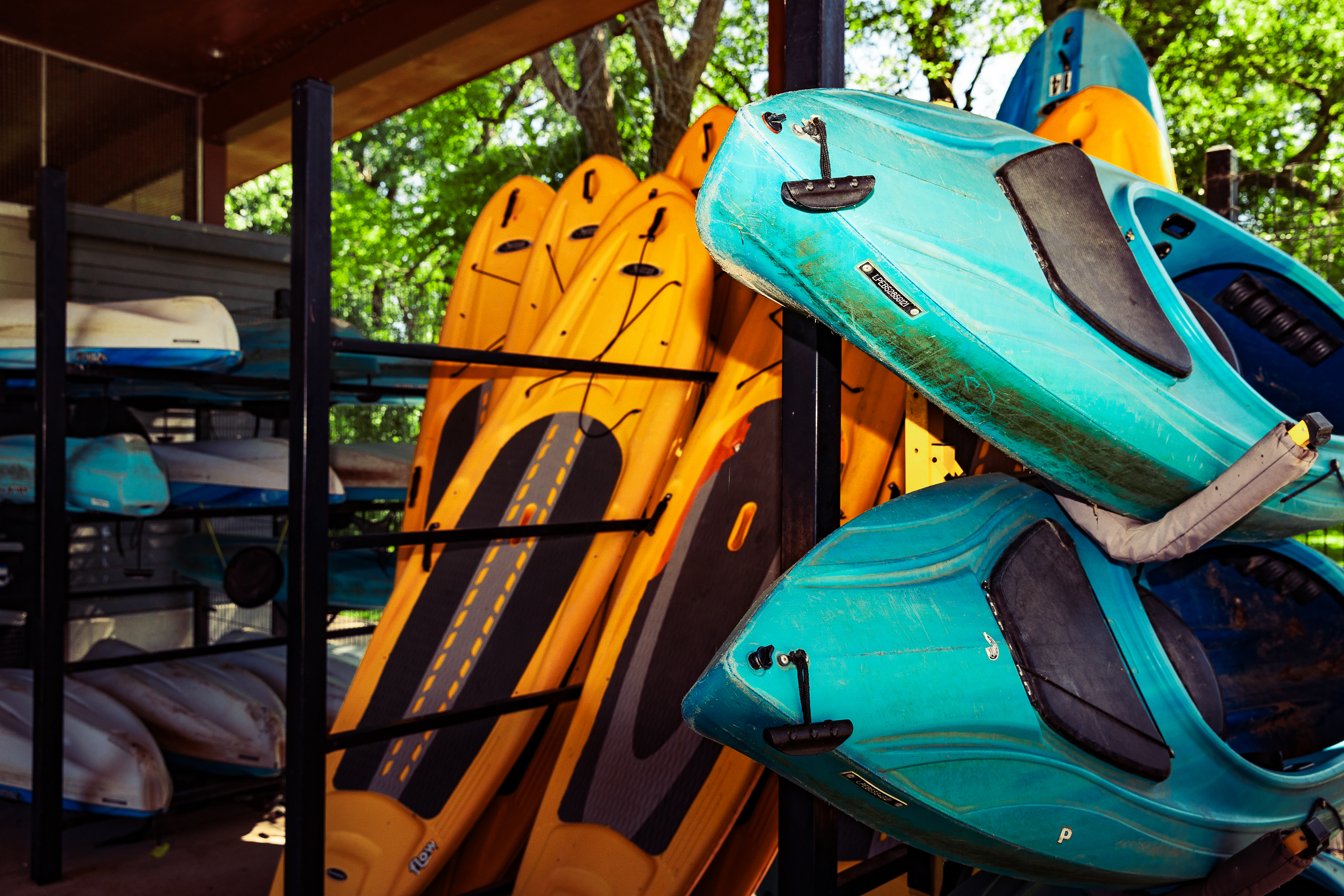 Stacked blue and orange kayaks on a rack outdoors, with sunlight filtering through trees and a turquoise kayak in the foreground.