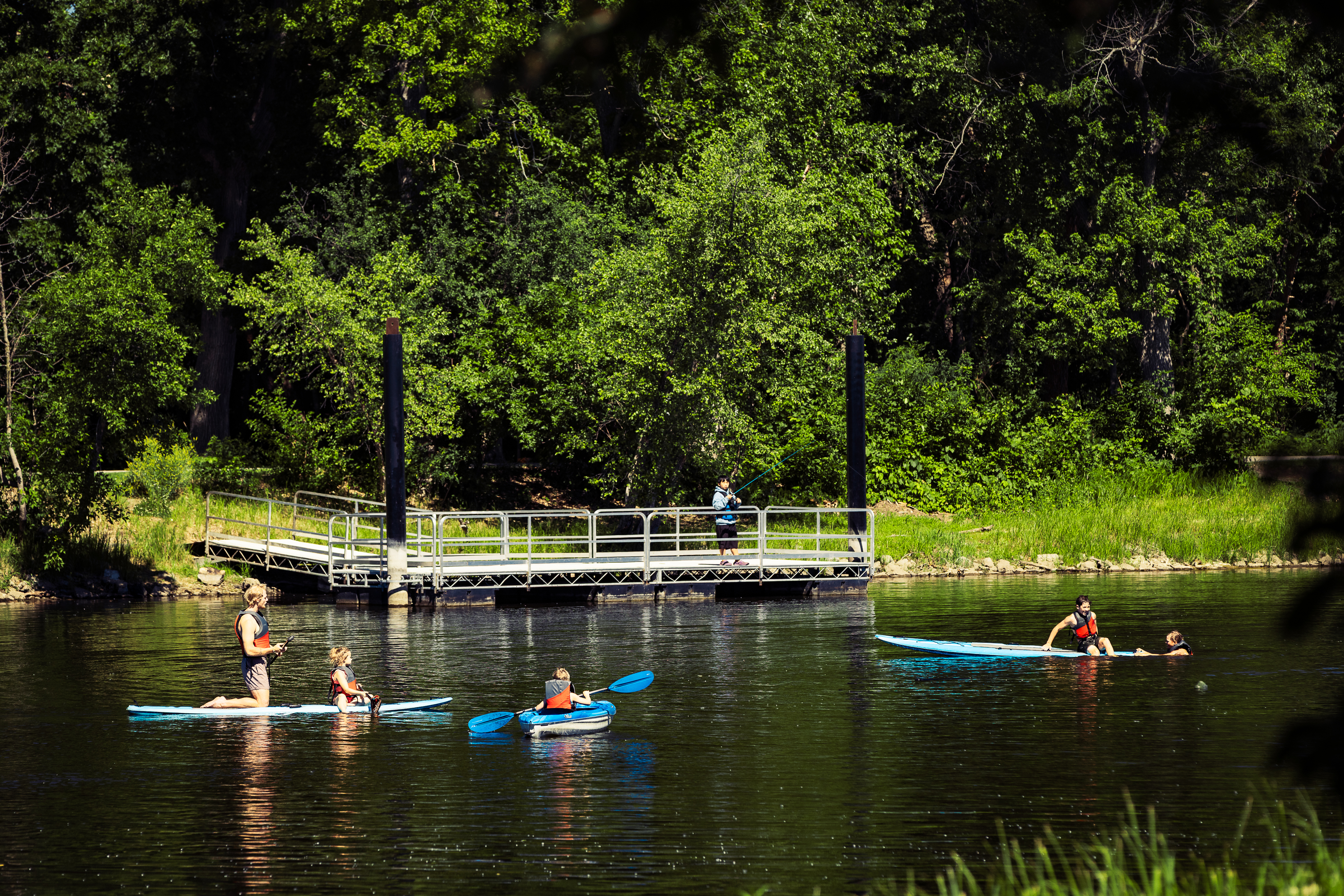 People paddle on a calm river with kayaks and a canoe near a floating dock, surrounded by dense green trees and dappled sunlight.